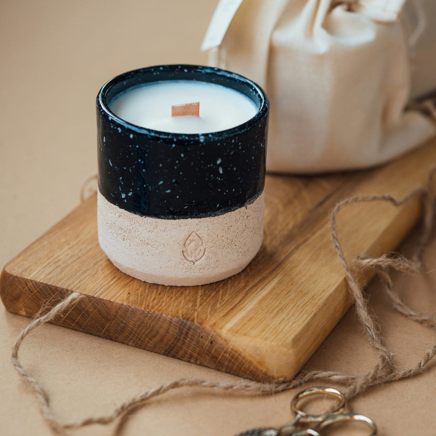 Sandalwood Classic Candle — A hand-poured candle in a speckled black ceramic cup with a wooden wick, beside a linen gift pouch