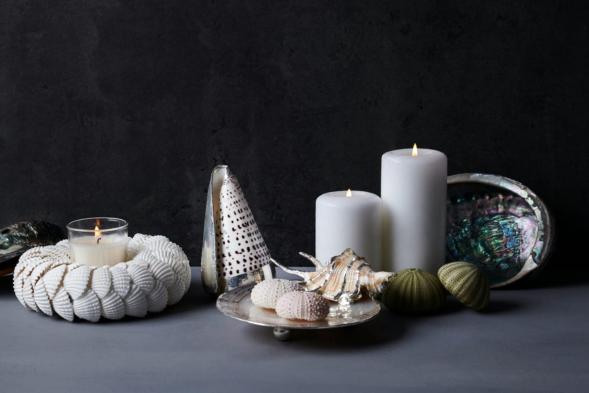 White pillar candles and a votive among seashells against a dark stone wall
