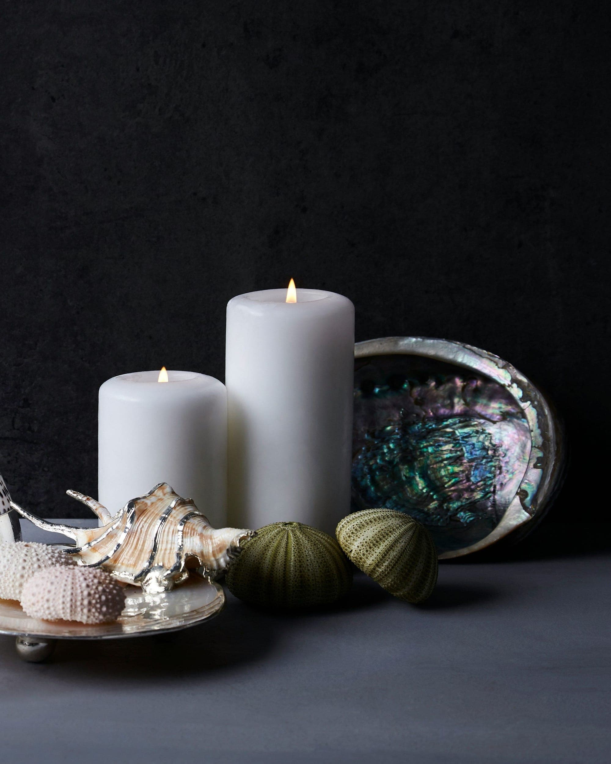 White pillar candles and a votive among seashells against a dark stone wall